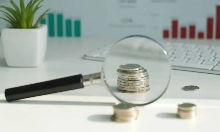 Magnifying glass focusing on stacked coins with financial charts in the background.