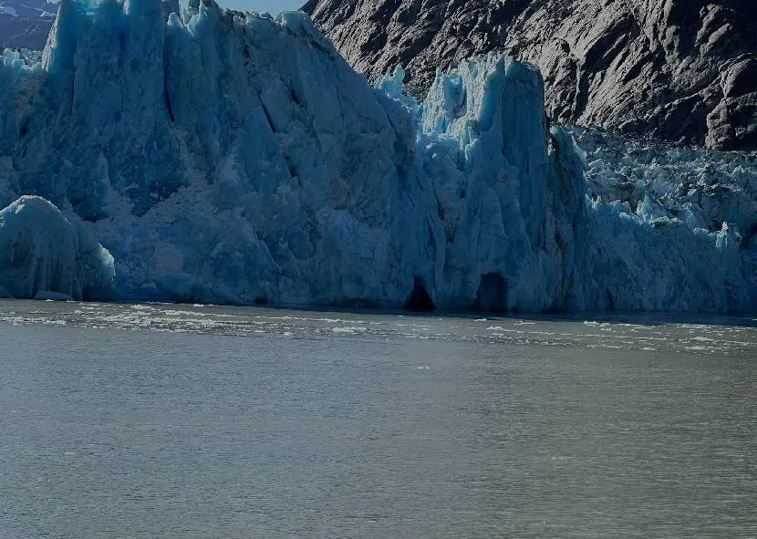 A massive blue glacier meets calm water against rocky terrain.