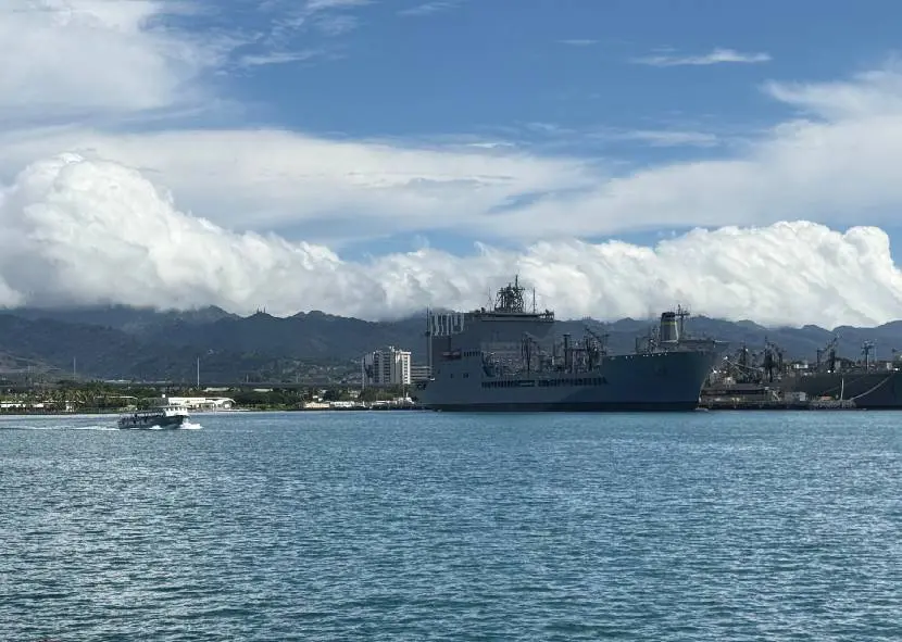 A large naval ship docked near a mountainous coastline under a partly cloudy sky.