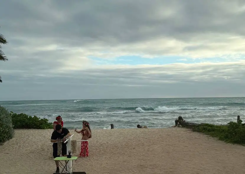A person and a child stand on a sandy beach near the ocean under a cloudy sky.